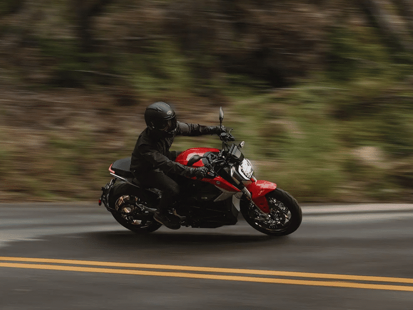 A person wearing a black helmet and dark clothing rides a red motorcycle at speed on a winding road, with blurred greenery in the background suggesting motion.