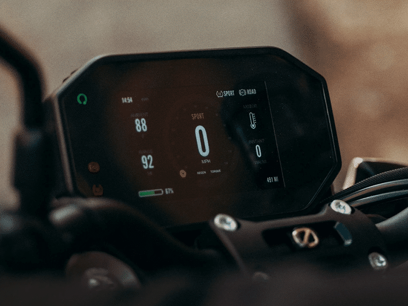 Close-up of a digital motorcycle dashboard displaying speed, battery level, ride mode, time, and temperature on a color screen. Handlebars and controls are partially visible in the foreground.