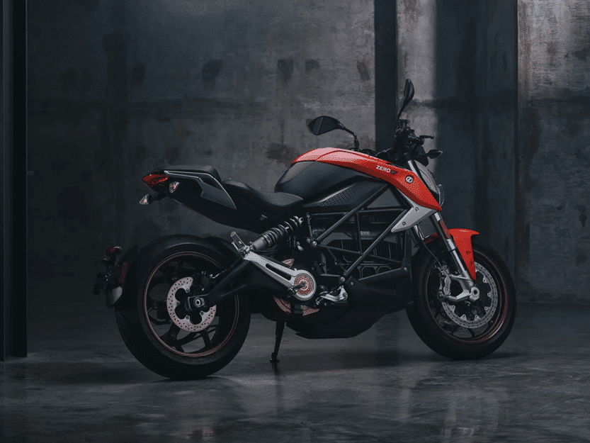 A sleek, red and black electric motorcycle is parked indoors against a dark, industrial concrete background, showcasing its modern design and exposed components.