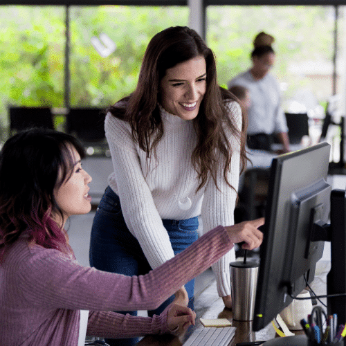 Two women in an office setting are looking at a computer screen together. One is seated and pointing at the monitor, while the other stands beside her, smiling and engaged. Other people and desks are blurred in the background.