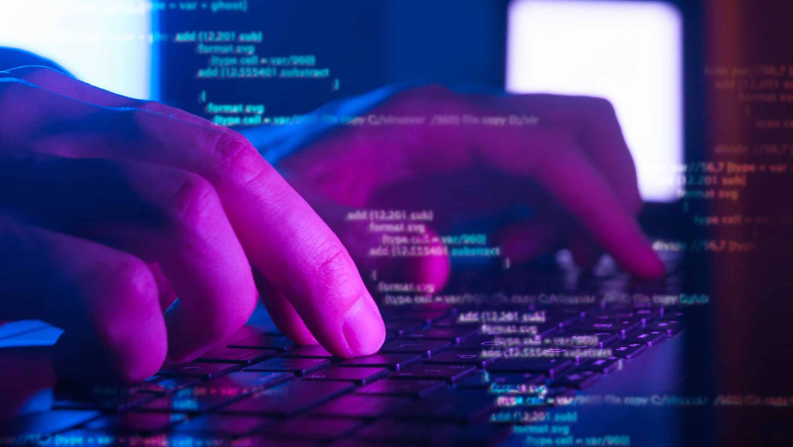 Close-up of hands typing on a laptop keyboard with lines of computer code overlaid, illuminated by purple and blue lighting, suggesting programming or cybersecurity work.