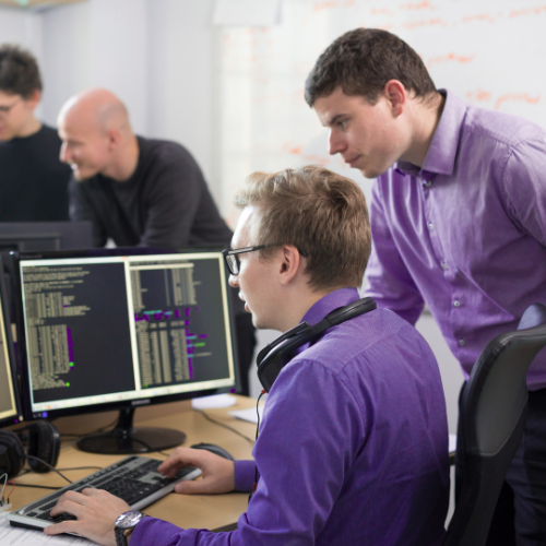 Two men in an office, one seated and one standing, look at computer screens displaying code. Both wear purple shirts and headphones are around the seated man’s neck. Two other people are blurred in the background.