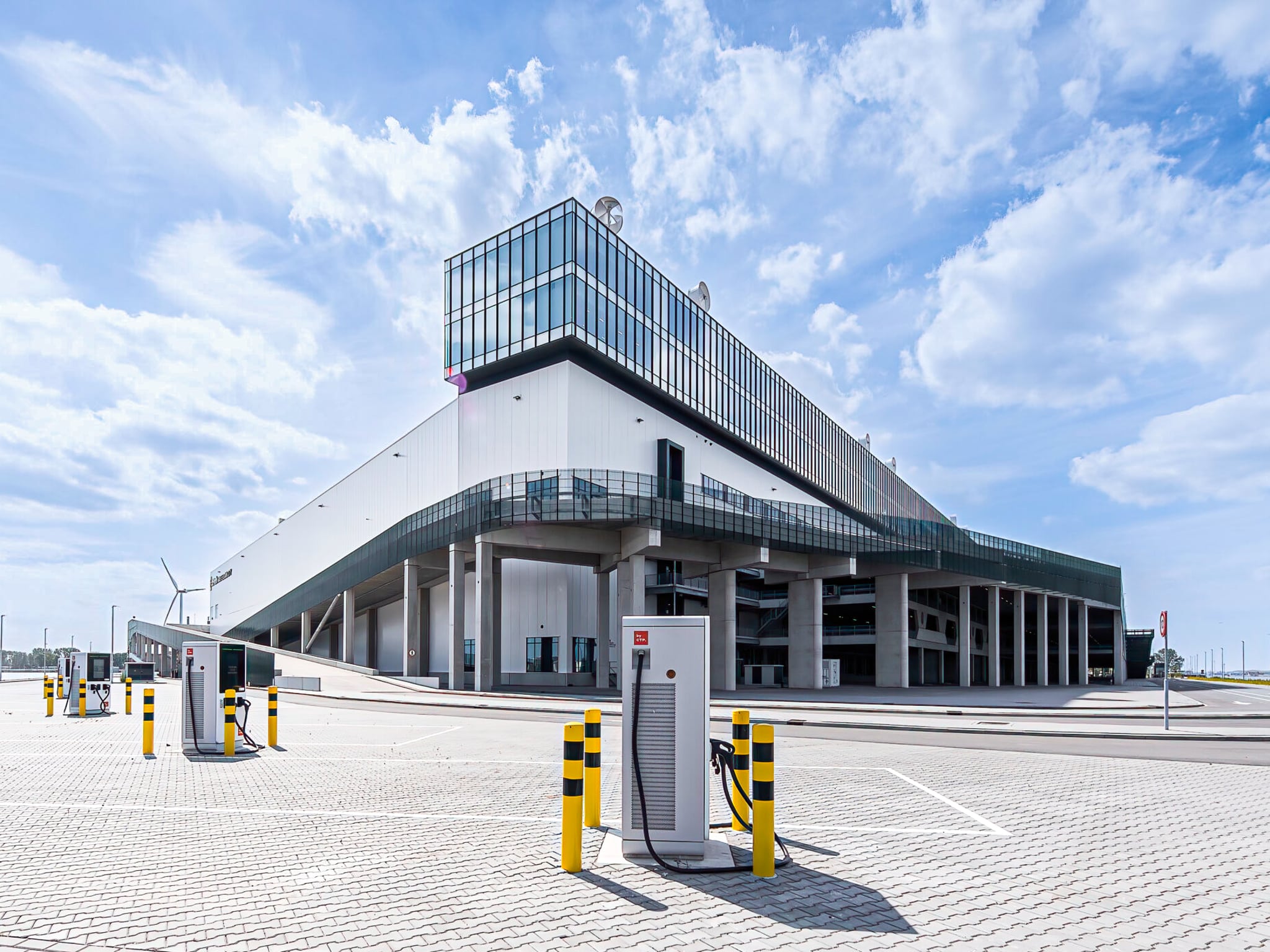 A modern, angular building with large glass windows and an elevated walkway, surrounded by an empty paved area and several electric vehicle charging stations under a partly cloudy sky.
