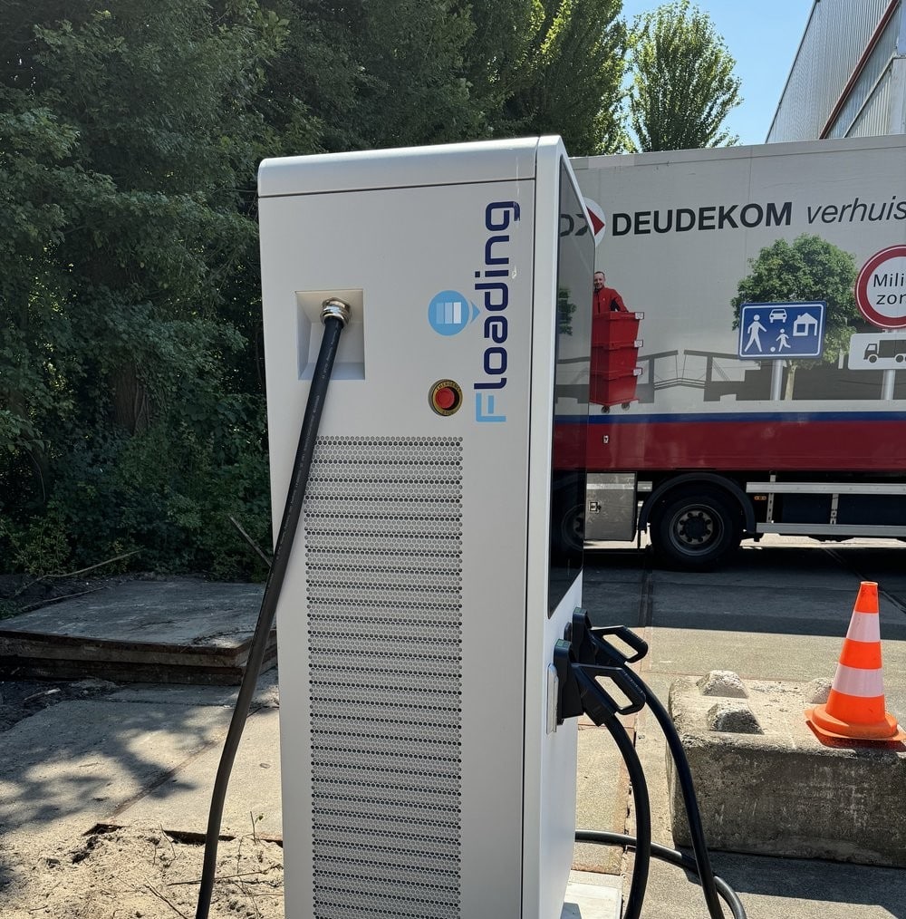 A white electric vehicle charging station labeled Flooading stands outdoors, with cables attached. A moving truck and orange traffic cone are visible in the background near trees and a building.