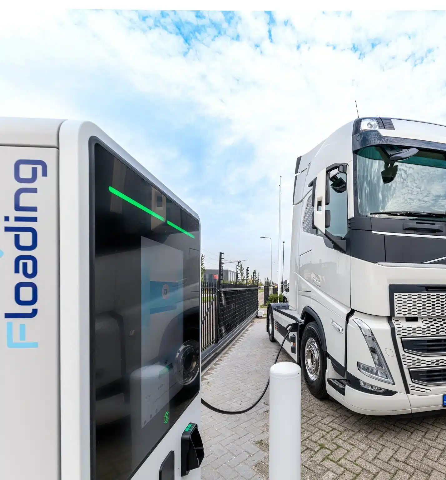 A white electric semi-truck is parked at a charging station, connected to the charger. The word loading is displayed vertically on the station. The background shows a cloudy sky and a paved area with fencing.