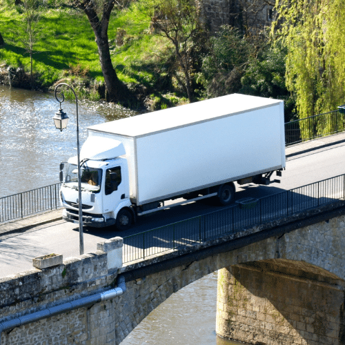 A white box truck drives over a stone bridge above a river, surrounded by greenery and trees on a sunny day.