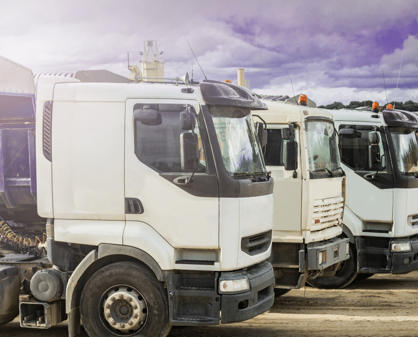 Several white semi trucks are parked in a row on a dirt lot under a cloudy sky.