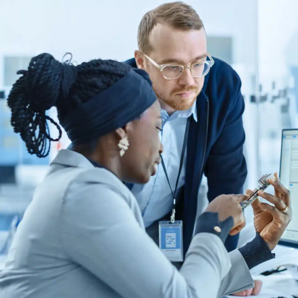 Two professionals in an office discuss a mechanical part while looking at a computer screen. The woman holds the part and gestures, while the man in glasses attentively observes, exploring the benefits of IoT Advisory Services.