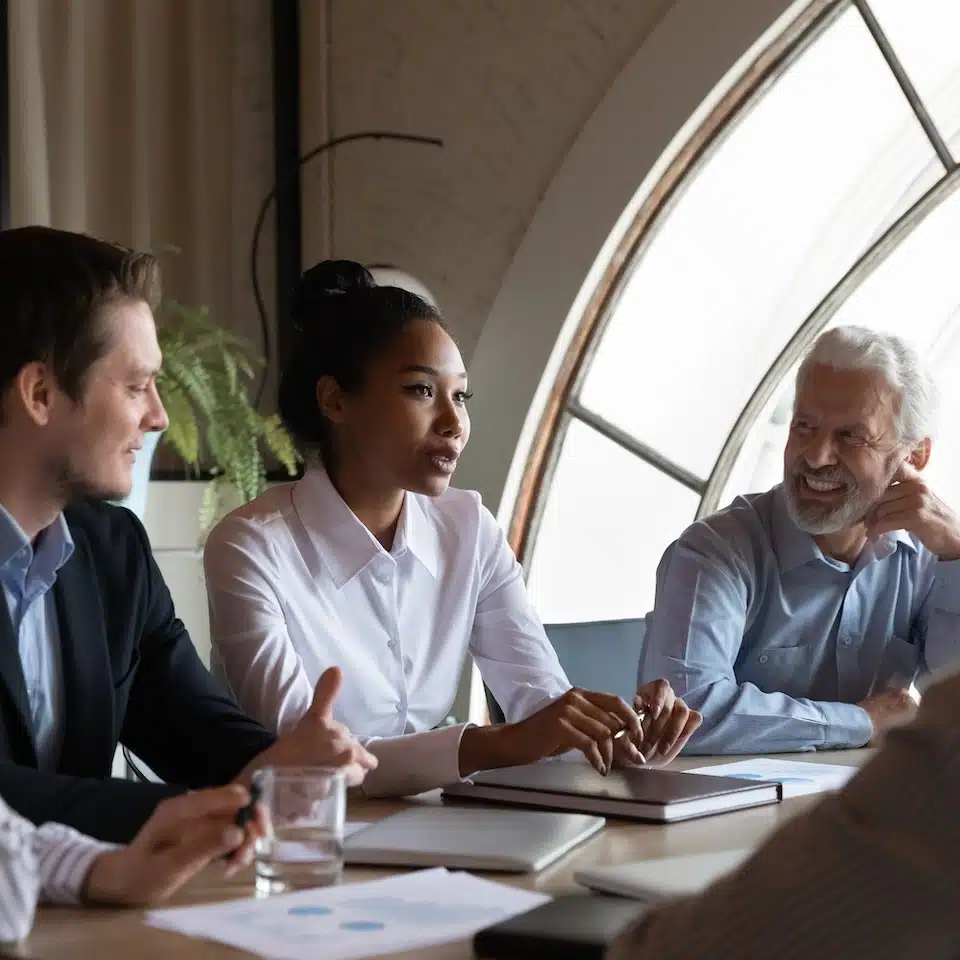 Three people sit at a conference table in a bright room having a discussion on IoT Advisory Services. A woman in a white shirt speaks while two men, one younger and one older, listen and smile. Papers and glasses of water are on the table.