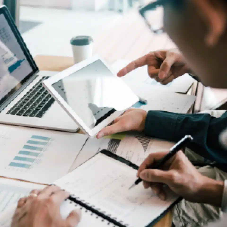 Two people review financial charts and graphs on paper, a laptop, and a tablet at a desk, discussing data and IoT Advisory Services while taking notes. A coffee cup sits in the background.