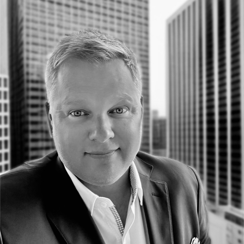 A man in a suit smiles at the camera, standing outdoors with tall office buildings in the background. The image is in black and white.