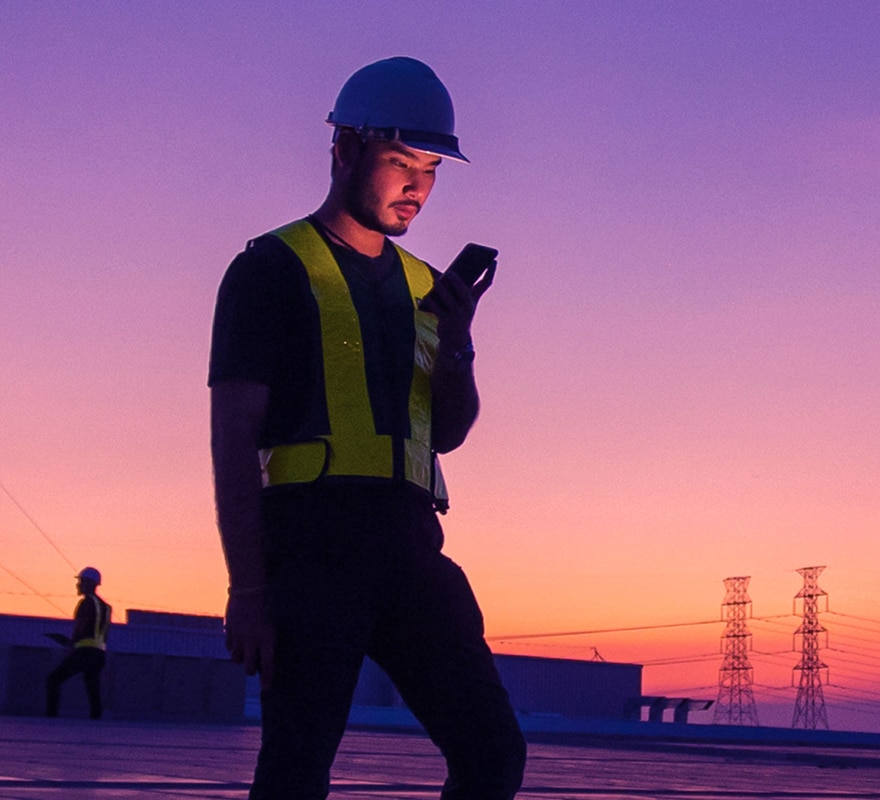 A construction worker wearing a helmet and reflective vest looks at his phone while standing outdoors at sunset, with another worker and power lines visible in the background.