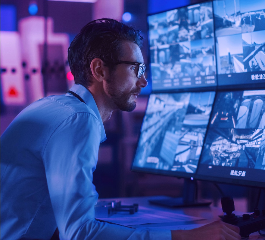 A man in glasses sits at a desk, monitoring multiple surveillance camera feeds on large screens in a dimly lit control room with blue and purple lighting.