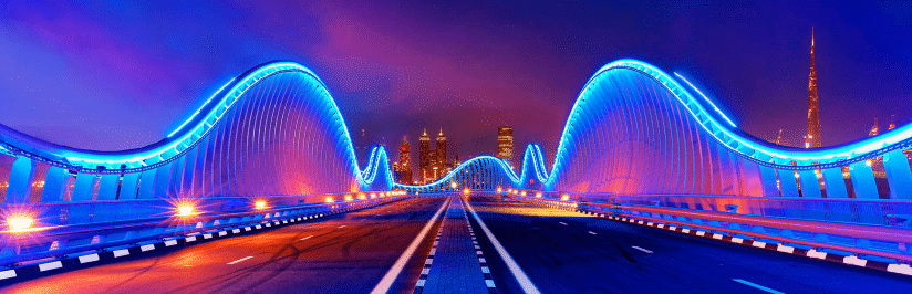 A modern bridge illuminated with blue lights curves under a vibrant evening sky, symbolizing the journey organizations face when deciding to build vs. buy an enterprise CMP. City skyscrapers and the iconic Burj Khalifa rise in the glowing background.
