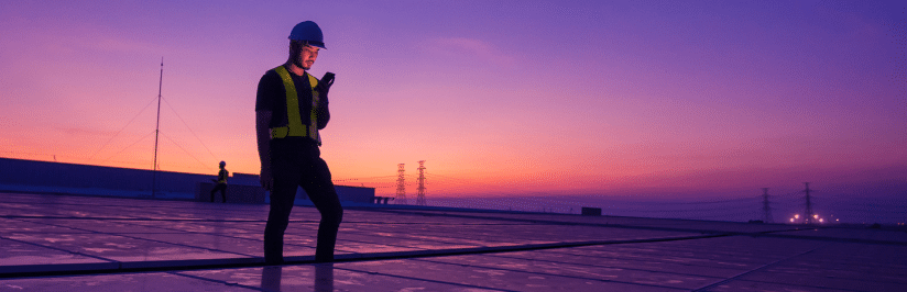 A worker wearing a hard hat and safety vest stands on a reflective rooftop at sunset, checking a mobile device—perhaps evaluating Build vs. Buy an Enterprise CMP—while another worker and power lines are visible in the background.