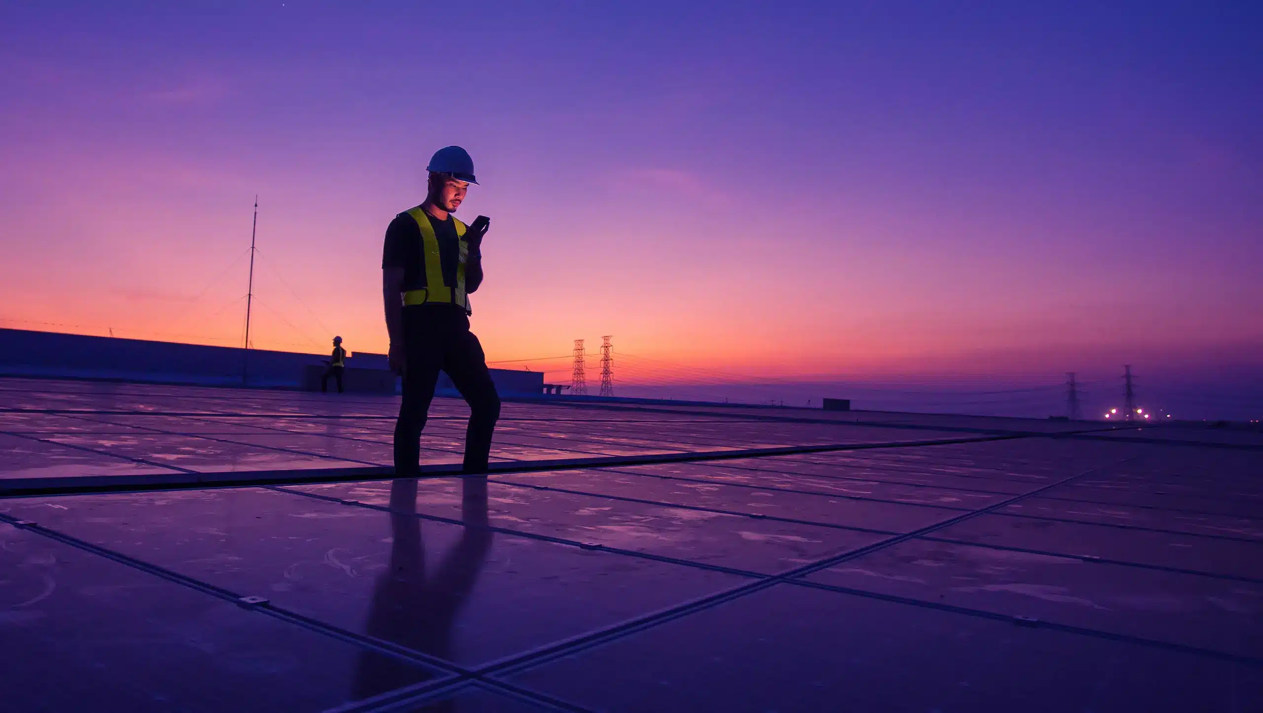 A worker wearing a hard hat and safety vest stands on a rooftop covered with solar panels at sunset, using a device connected to the Hera 600 IoT router. The sky glows purple and orange, with power lines visible in the background.