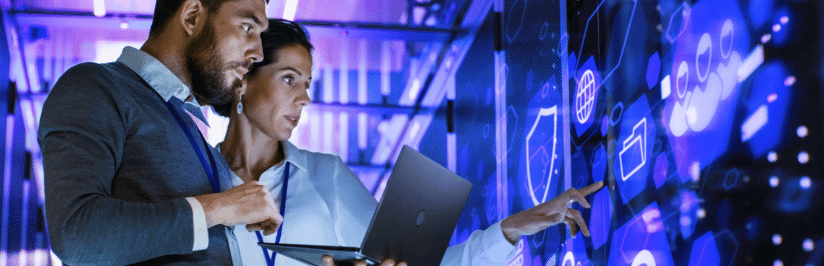 Two people in business attire examine a digital screen displaying data and security icons related to FWA in a high-tech environment. One holds a laptop while the other points to the display, both focused on fixed wireless access information.