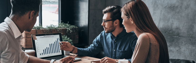 Three people sit at a table in an office, discussing a bar graph on a laptop screen showing 5G FWA trends. One man points at the graph while the others listen attentively. Coffee cups and notebooks are on the table.