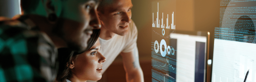 Three people closely examine computer screens displaying data visualizations and charts in a dimly lit room, suggesting collaborative analysis or teamwork in a tech or office environment.