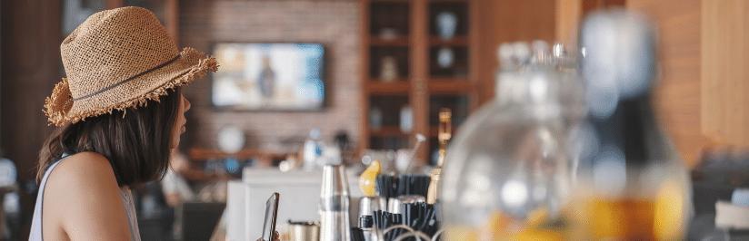 A woman wearing a straw hat sits at a counter in a cafe or bar, holding her phone—perhaps browsing UK managed fixed wireless access for Hospitality. The foreground is blurred with bottles and utensils, shelves and a TV visible behind her.